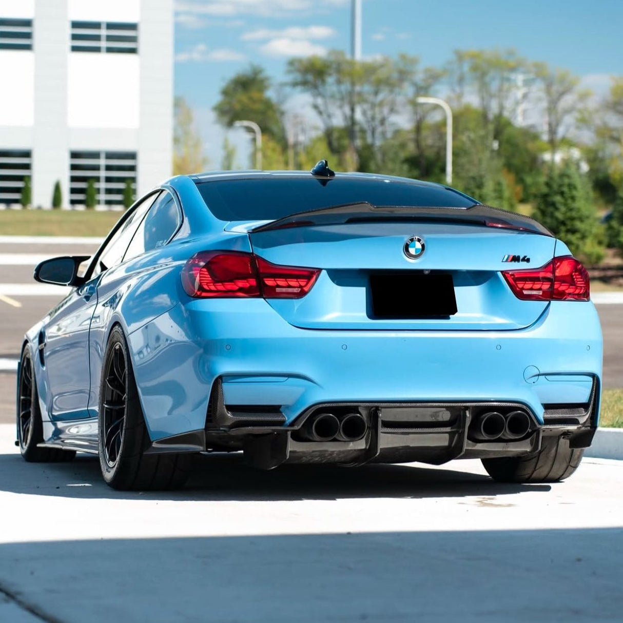 A blue BMW M4 car is parked on a paved surface with a modern building and greenery in the background.