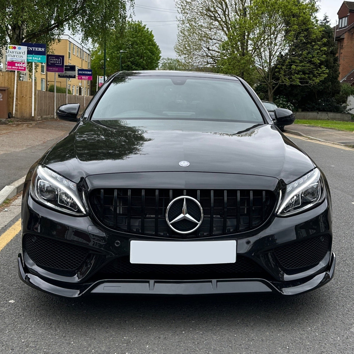 A sleek black car, parked on a suburban street, features a prominent Mercedes-Benz logo on the grille. Surrounding are trees and buildings with real estate signs visible in the background.