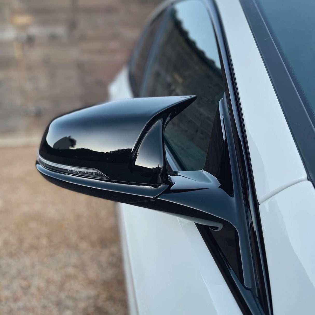 A sleek black car side mirror reflects the sky, attached to a white vehicle. The background is a blurred, neutral-toned wall.