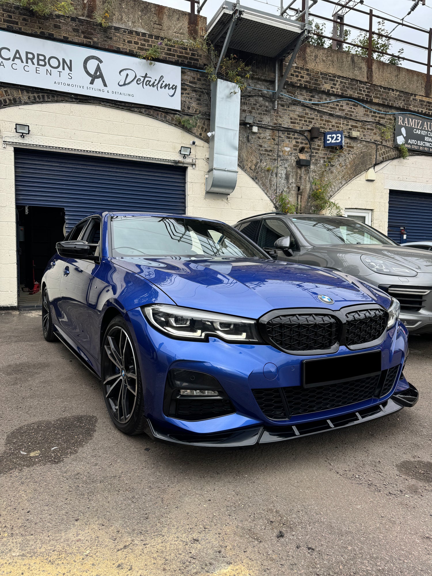 A blue car, parked outside an automotive detailing shop, features sleek design elements. Signs read "CARBON ACCENTS Detailing" and "RAMIZ AUTO," emphasizing automotive styling services. A wall with greenery surrounds the lot.