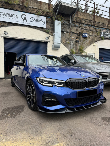 A blue car, parked outside an automotive detailing shop, features sleek design elements. Signs read "CARBON ACCENTS Detailing" and "RAMIZ AUTO," emphasizing automotive styling services. A wall with greenery surrounds the lot.