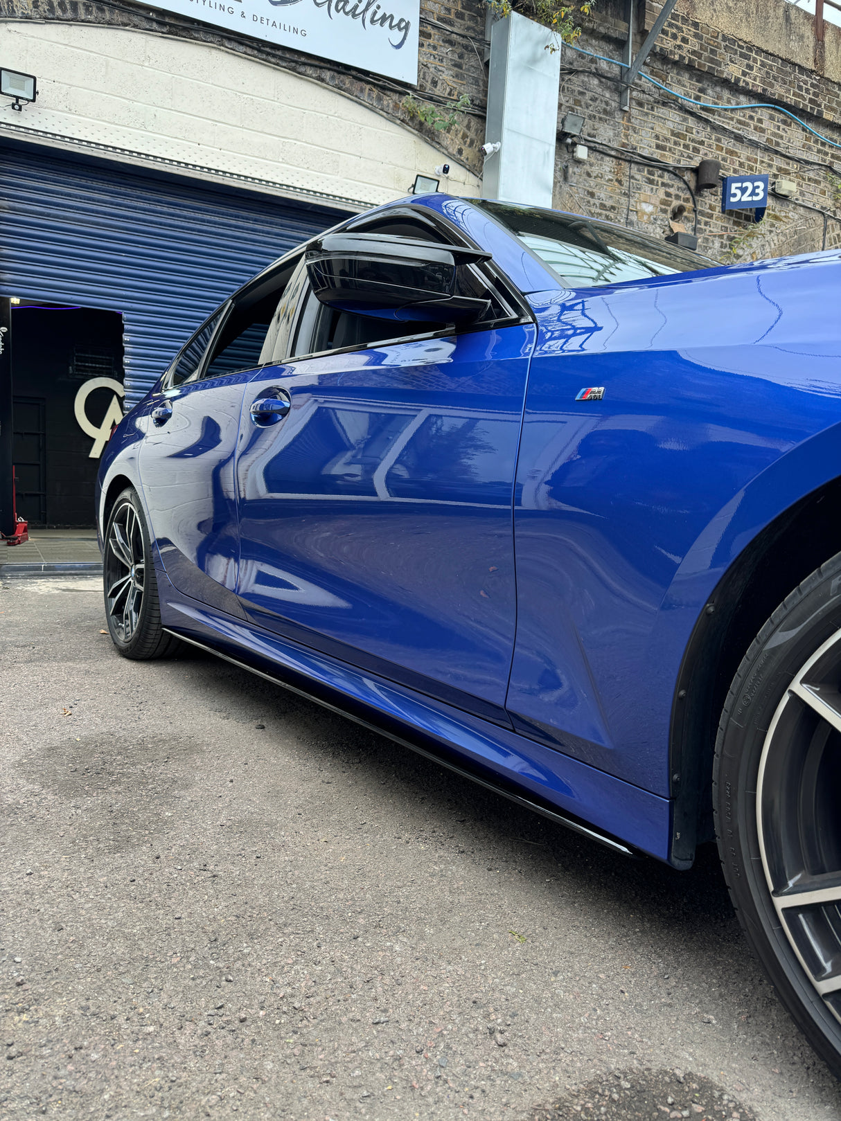 A blue car, parked with its window slightly open, is situated outside a garage. A sign reads "Reviving & Detailing." A building number "523" is visible on a brick wall.