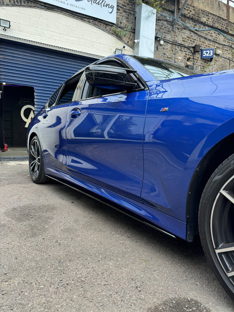 A blue car, parked with its window slightly open, is situated outside a garage. A sign reads "Reviving & Detailing." A building number "523" is visible on a brick wall.
