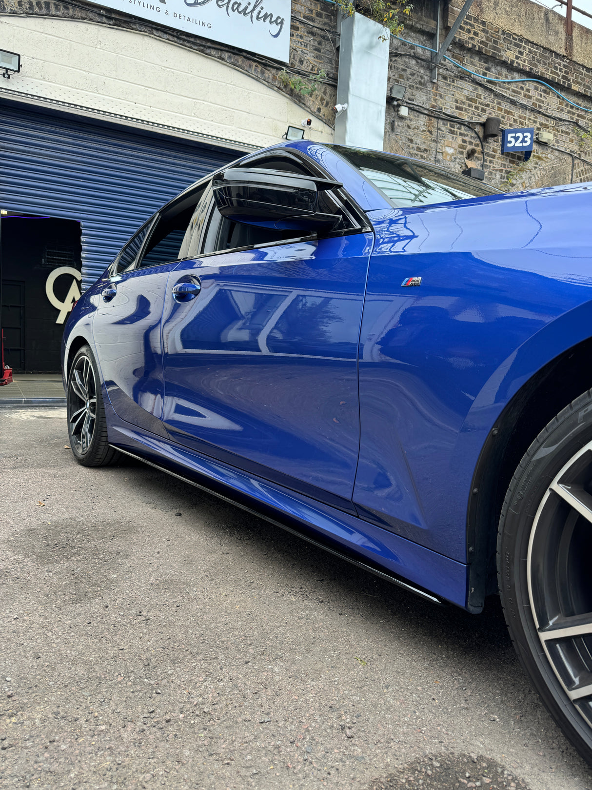 A blue car parked with its window slightly open, located outside a detailing garage beneath a brick archway. The garage has a sign reading “Styling & Detailing” and a number “523.”