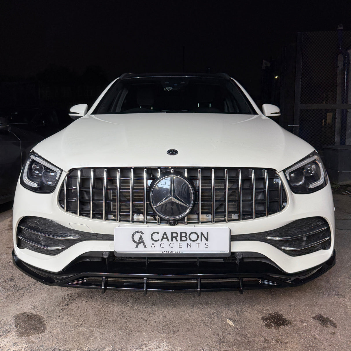 A white SUV is parked on a concrete surface in front of a dark backdrop. The front grille features a prominent Mercedes emblem. The license plate reads "CARBON ACCENTS".