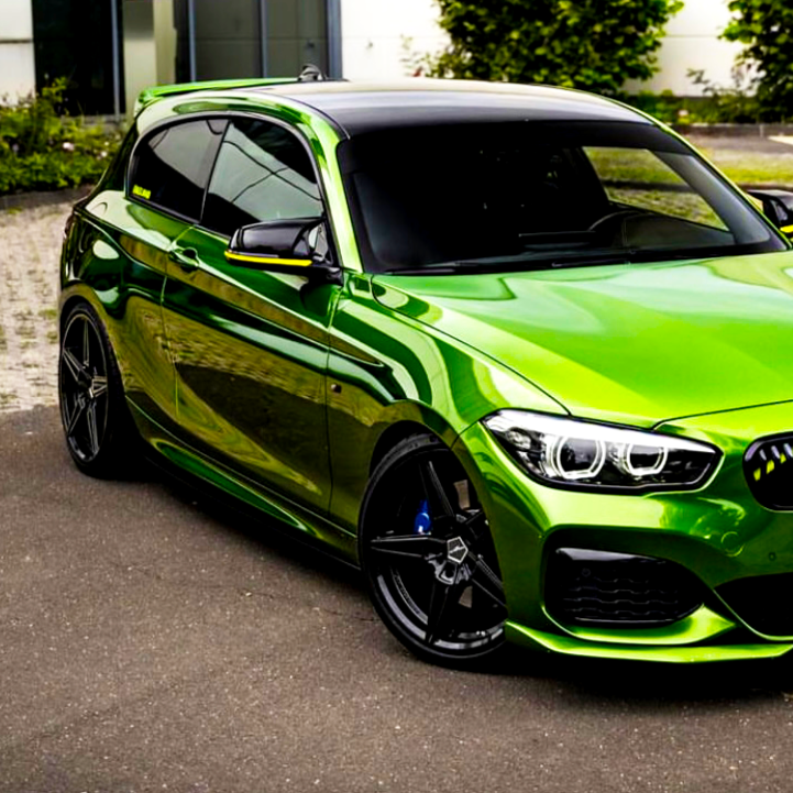 A vibrant green car parked on pavement, reflecting sunlight, surrounded by greenery and a building with large windows. The car has black wheels and tinted windows.