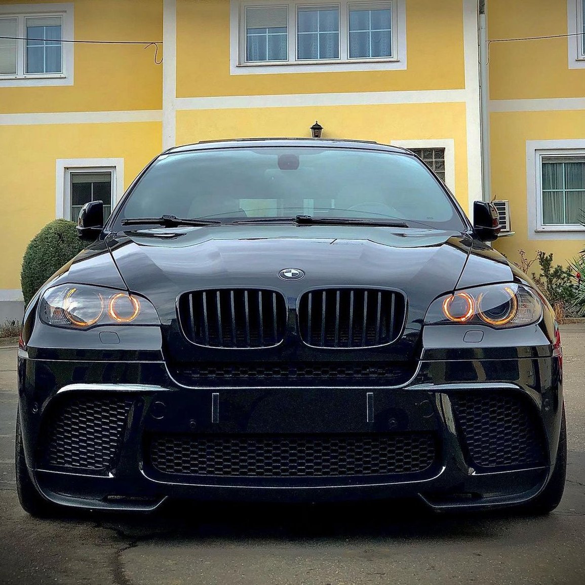 A black BMW with illuminated circular headlights is parked in front of a yellow building with white trim and windows, creating a striking contrast between the car and its surroundings.