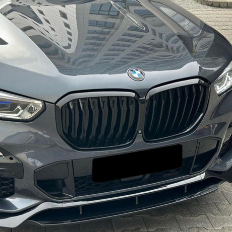 A shiny gray BMW car with a prominent black grille and blue-accented headlights is parked on interlocking paving stones. A building is reflected on the hood.