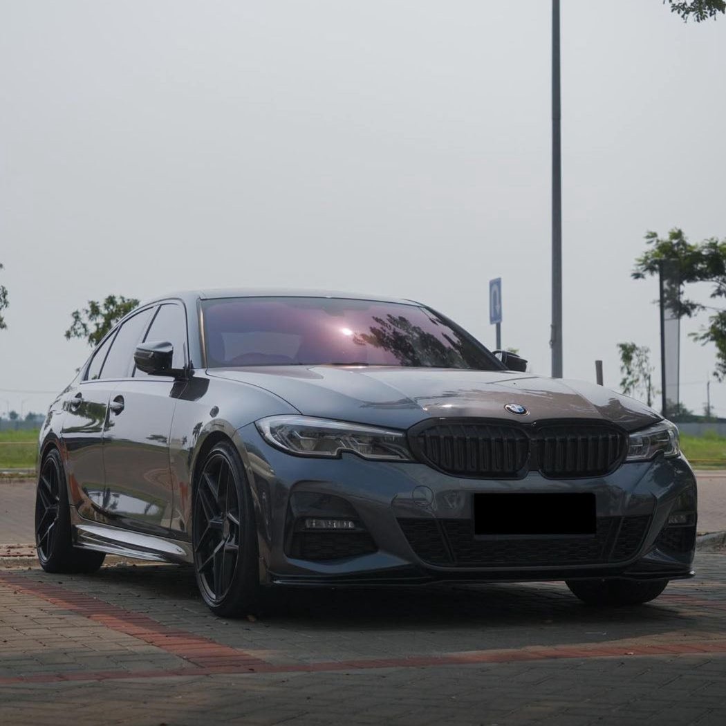 A sleek, silver BMW sedan is parked on a paved area, under a cloudy sky, surrounded by sparse trees and a street sign.