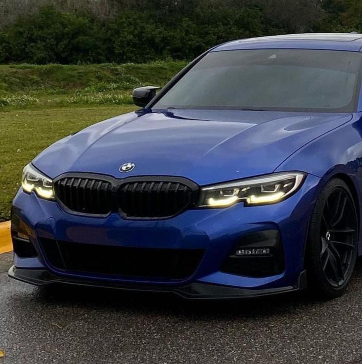A blue BMW with its headlights on is parked on a wet road, surrounded by grass and trees in the background.