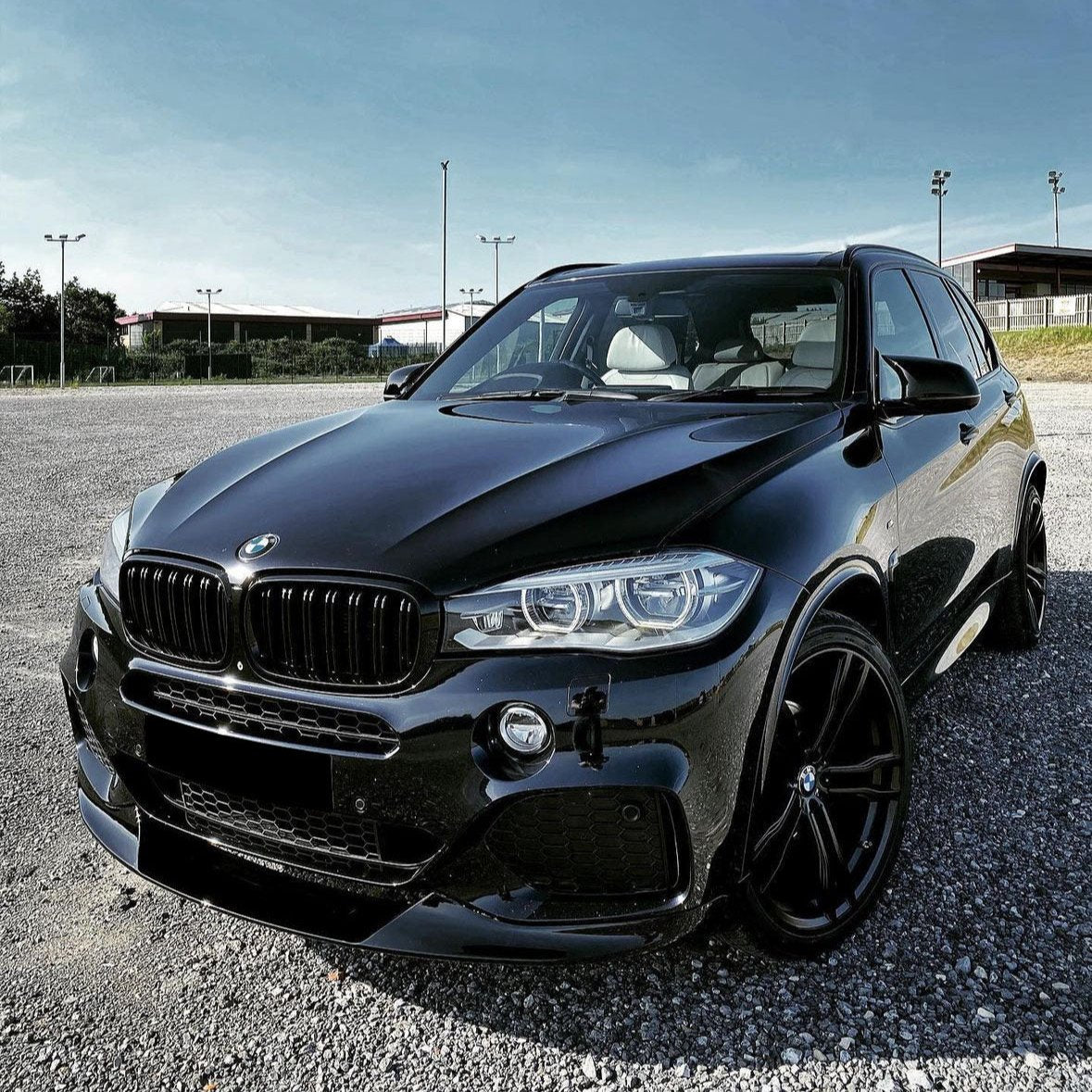 A sleek black BMW SUV is parked on a gravel lot. It sits under a clear blue sky, near empty sports fields with pole lights.