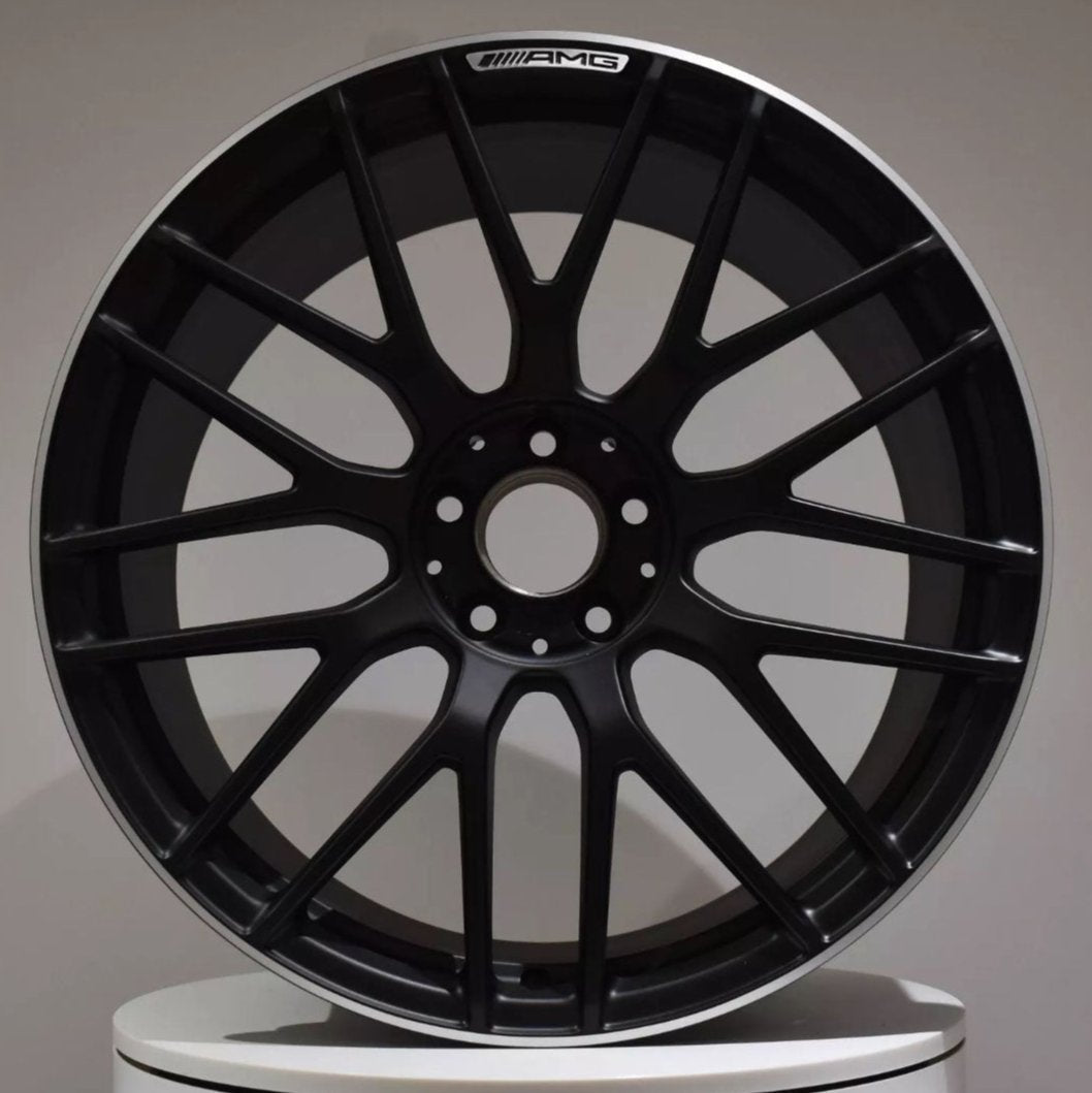 A black, multi-spoke AMG car rim is displayed upright against a neutral background, resting on a white surface.