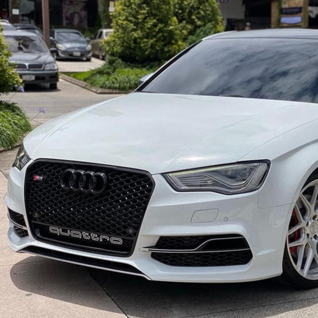 A white Audi car with a prominent black grille is parked on a concrete surface. The word "quattro" is visible on the grille. Trees and other cars are in the background.