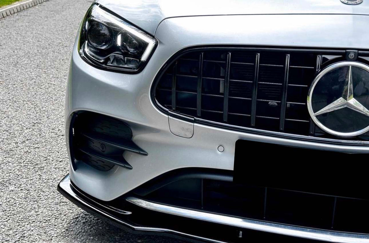 A silver car's front grille displays a prominent Mercedes-Benz emblem, parked on a gravel road with grass bordering the pavement.