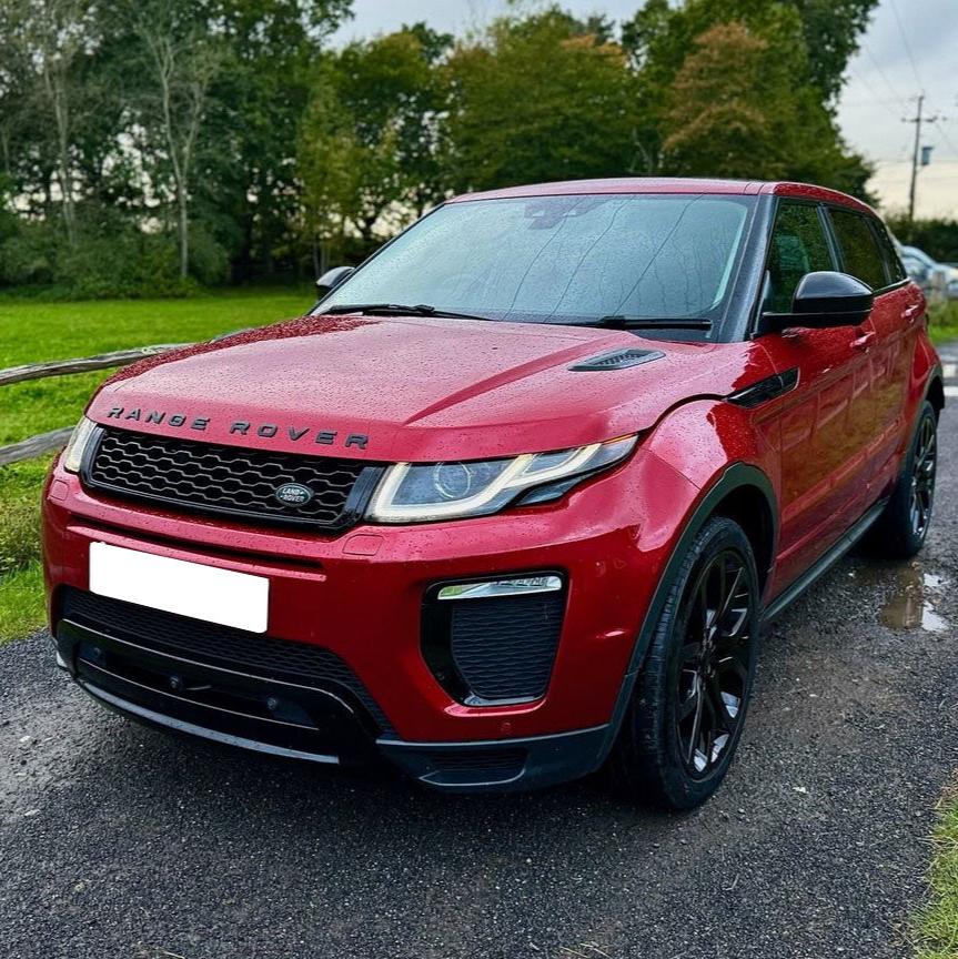 Red Range Rover parked, covered in raindrops. Situated on a wet, paved road beside a green field with trees. "Range Rover" is written on the front.