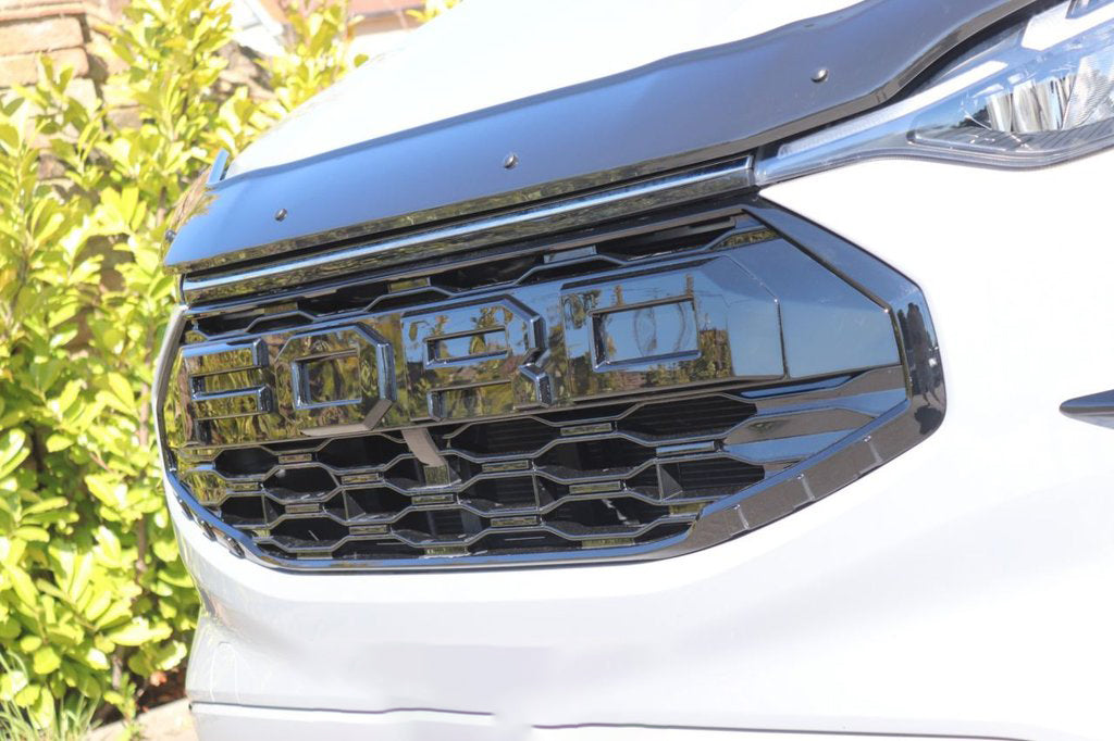 A white vehicle's front grille, featuring the letters "FORD," is prominently displayed. The vehicle is parked outdoors, surrounded by green foliage and sunlight.