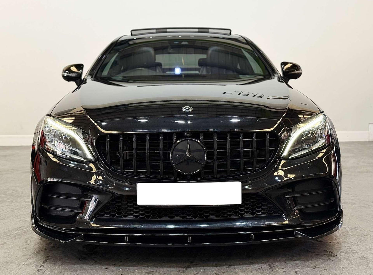 A black luxury sedan with illuminated headlights is parked on a concrete floor in a white-walled room. The license plate is blank, and a Mercedes-Benz emblem is visible.