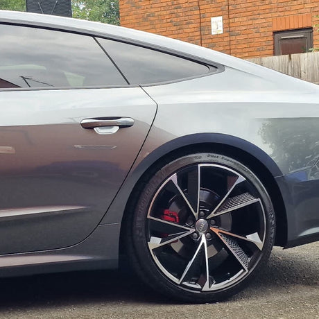 A sleek silver car with tinted windows is parked beside a brick building. The focus is on the car's rear wheel, which features a stylized rim design and red brake caliper.