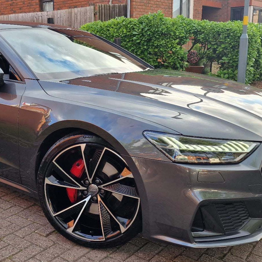 A sleek gray car is parked on a paved driveway, showcasing its polished body, distinctive alloy wheel, and illuminated headlights against a background of green hedges and brick buildings.