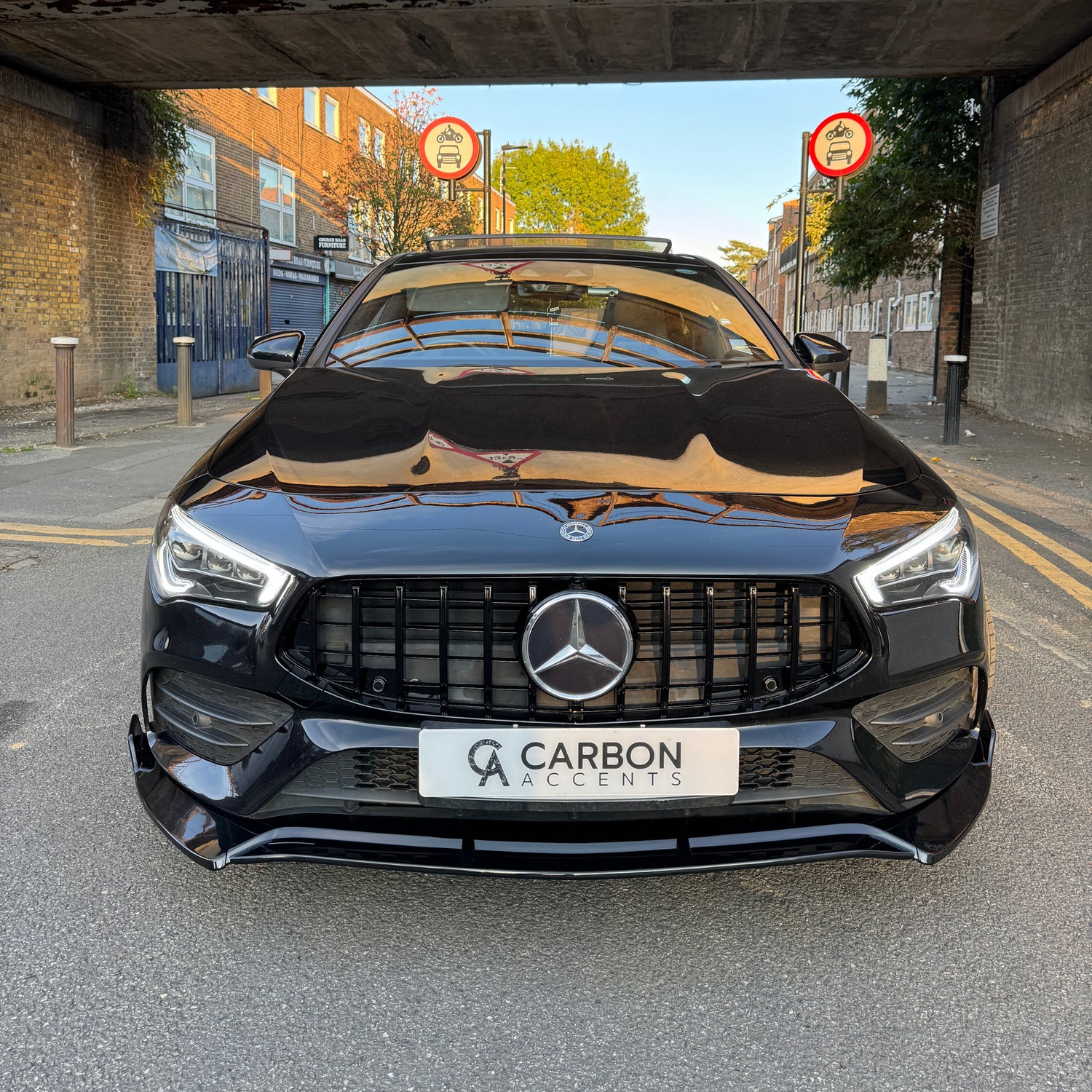 A black car with a Mercedes emblem is parked under a bridge. Reflective windows and "CARBON ACCENTS" text on the plate are visible. Road signs and urban buildings are in the background.