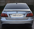 The image shows the rear of a silver Mercedes-Benz E63 AMG with illuminated tail lights, parked on a brick driveway. The license plate reads "CARBON ACCENTS." The background features a wooden fence.