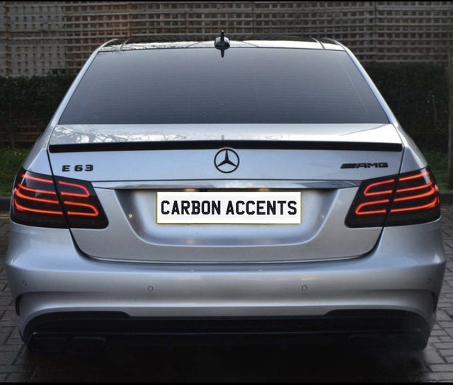 The image shows the rear of a silver Mercedes-Benz E63 AMG with illuminated tail lights, parked on a brick driveway. The license plate reads "CARBON ACCENTS." The background features a wooden fence.