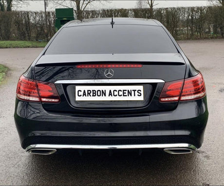 Car with black paint and a "CARBON ACCENTS" license plate remains stationary in a parking area. Background includes leafless trees and a green container.