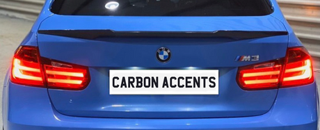 A blue car with illuminated red taillights is parked in an indoor setting. The license plate reads "CARBON ACCENTS." The car has a BMW badge and M3 emblem on the back.