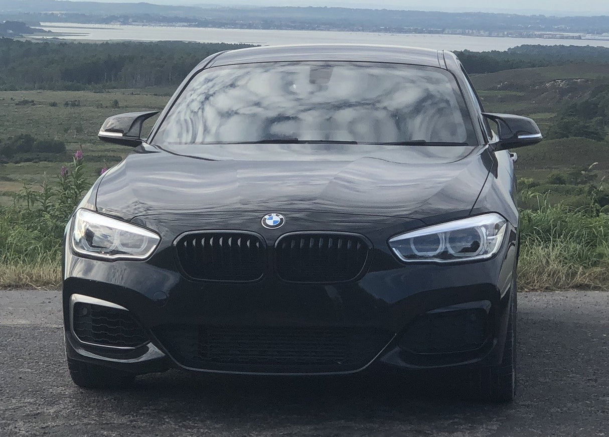 A black BMW car is parked on a road, facing forward, with rolling hills and a body of water in the distant background.