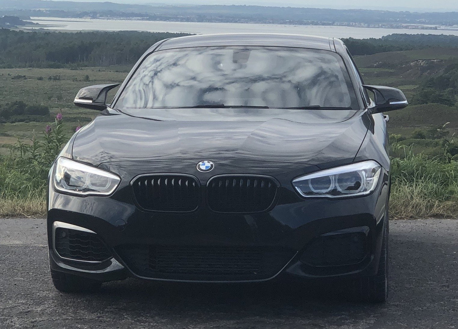 A black BMW car is parked on a road, facing forward, with rolling hills and a body of water in the distant background.