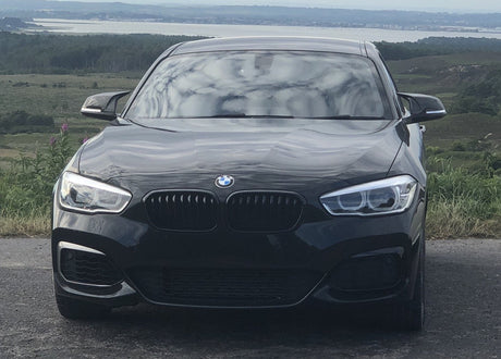 A black BMW car is parked on a road, facing forward, with rolling hills and a body of water in the distant background.