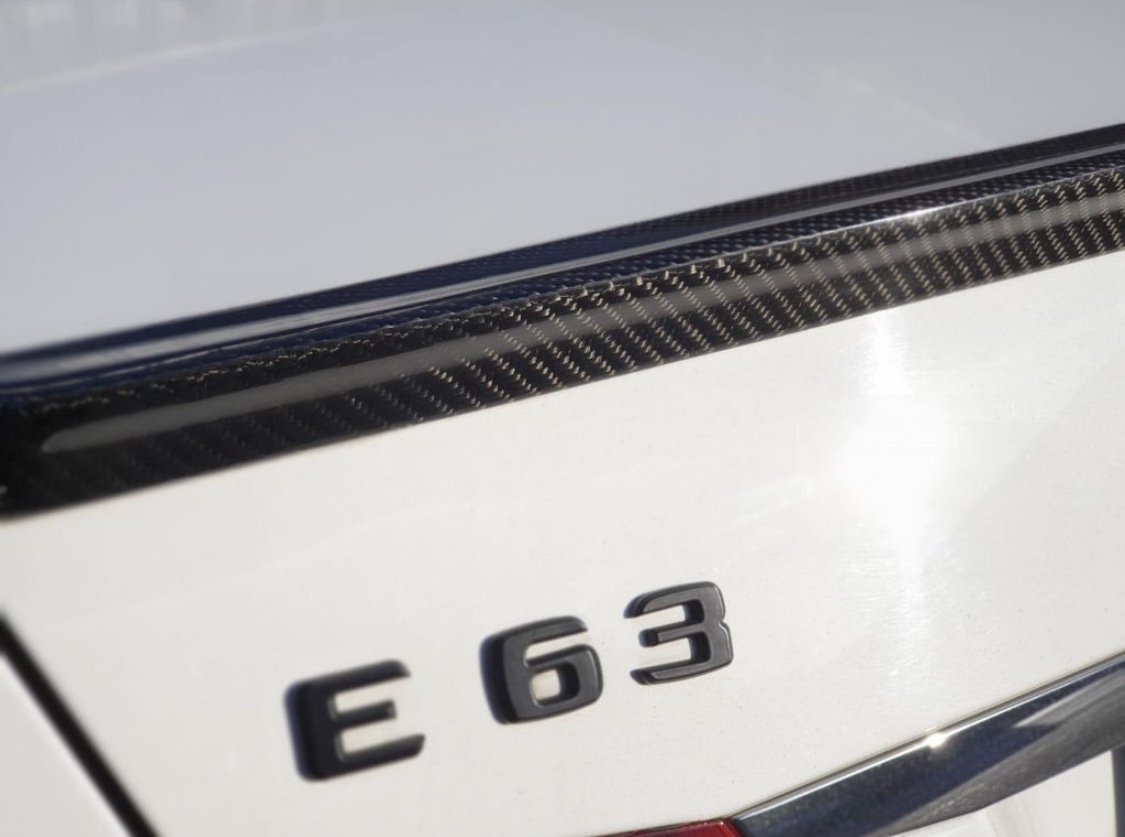 The image shows the rear emblem "E 63" on a white car, featuring a carbon fiber detail on the trunk lid in bright sunlight.