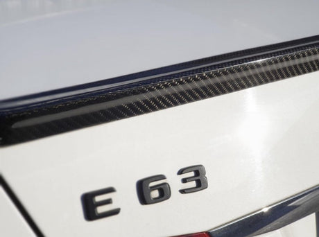 The image shows the rear emblem "E 63" on a white car, featuring a carbon fiber detail on the trunk lid in bright sunlight.
