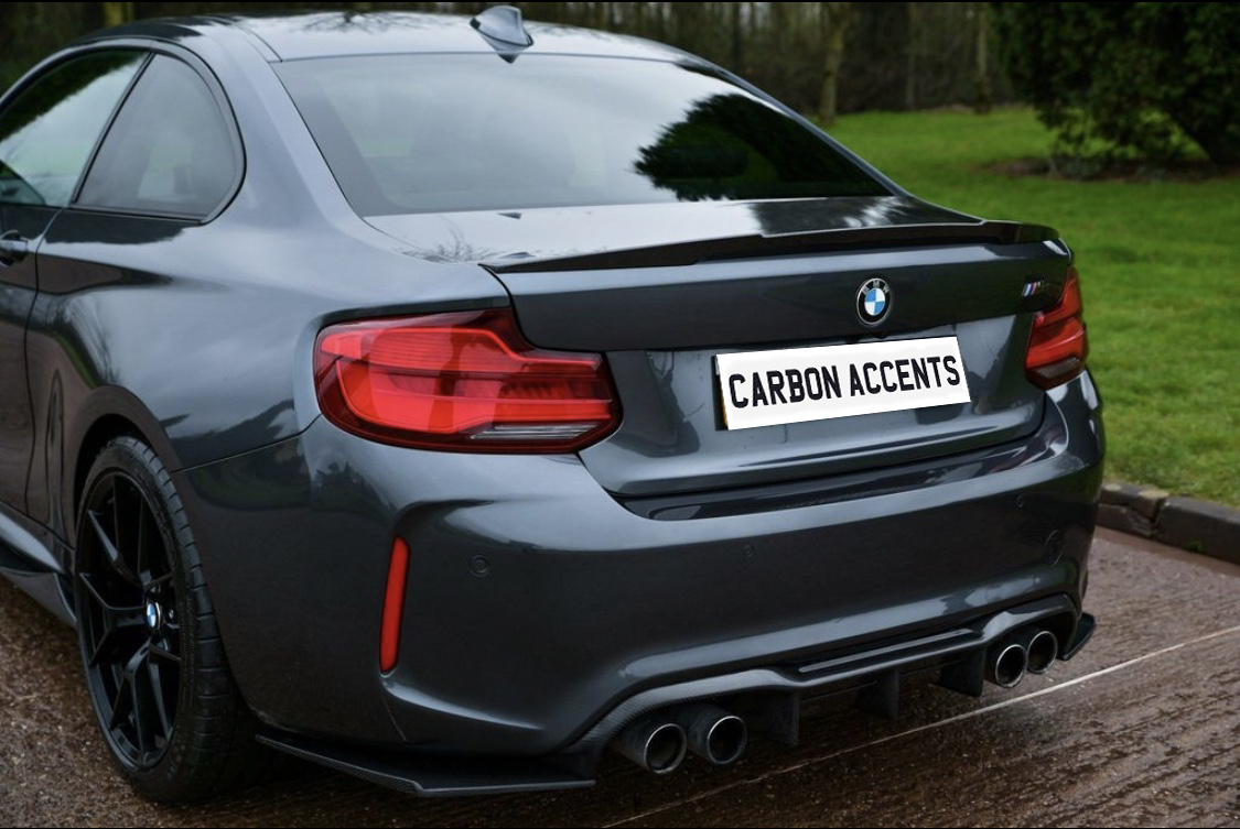 A dark gray BMW sports car is parked on a wet road, featuring a dual exhaust and distinctive rear lights, with a license plate reading "CARBON ACCENTS" in a green, grassy environment.