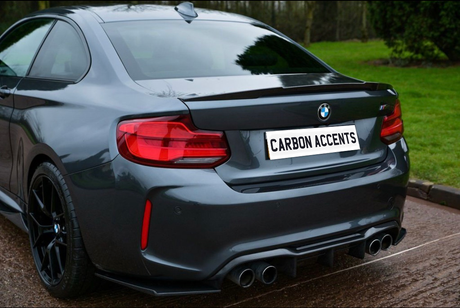 A dark gray BMW sports car is parked on a wet road, featuring a dual exhaust and distinctive rear lights, with a license plate reading "CARBON ACCENTS" in a green, grassy environment.