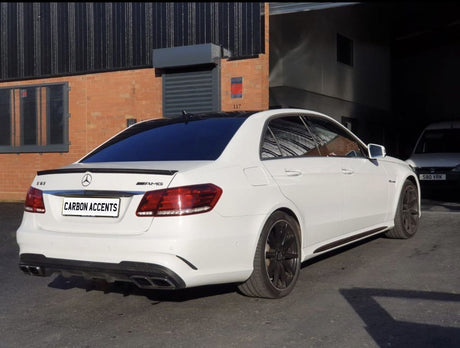 A white Mercedes-Benz E63 AMG is parked on a paved area outside an industrial building with orange brick walls. The text "CARBON ACCENTS" is on the license plate.