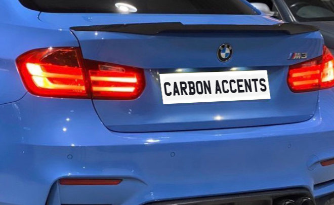 A blue BMW M3 with illuminated rear lights is parked. The license plate reads "CARBON ACCENTS," and a carbon fiber spoiler is visible on its trunk. Another car is adjacent.