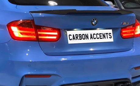 A blue BMW M3 with illuminated rear lights is parked. The license plate reads "CARBON ACCENTS," and a carbon fiber spoiler is visible on its trunk. Another car is adjacent.