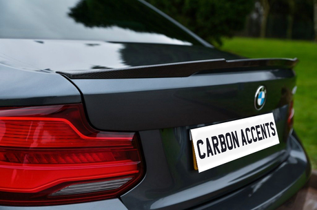 A car's rear shows a sleek spoiler and a visible logo, with text reading "CARBON ACCENTS" on the license plate. It's parked on a driveway beside green grass.