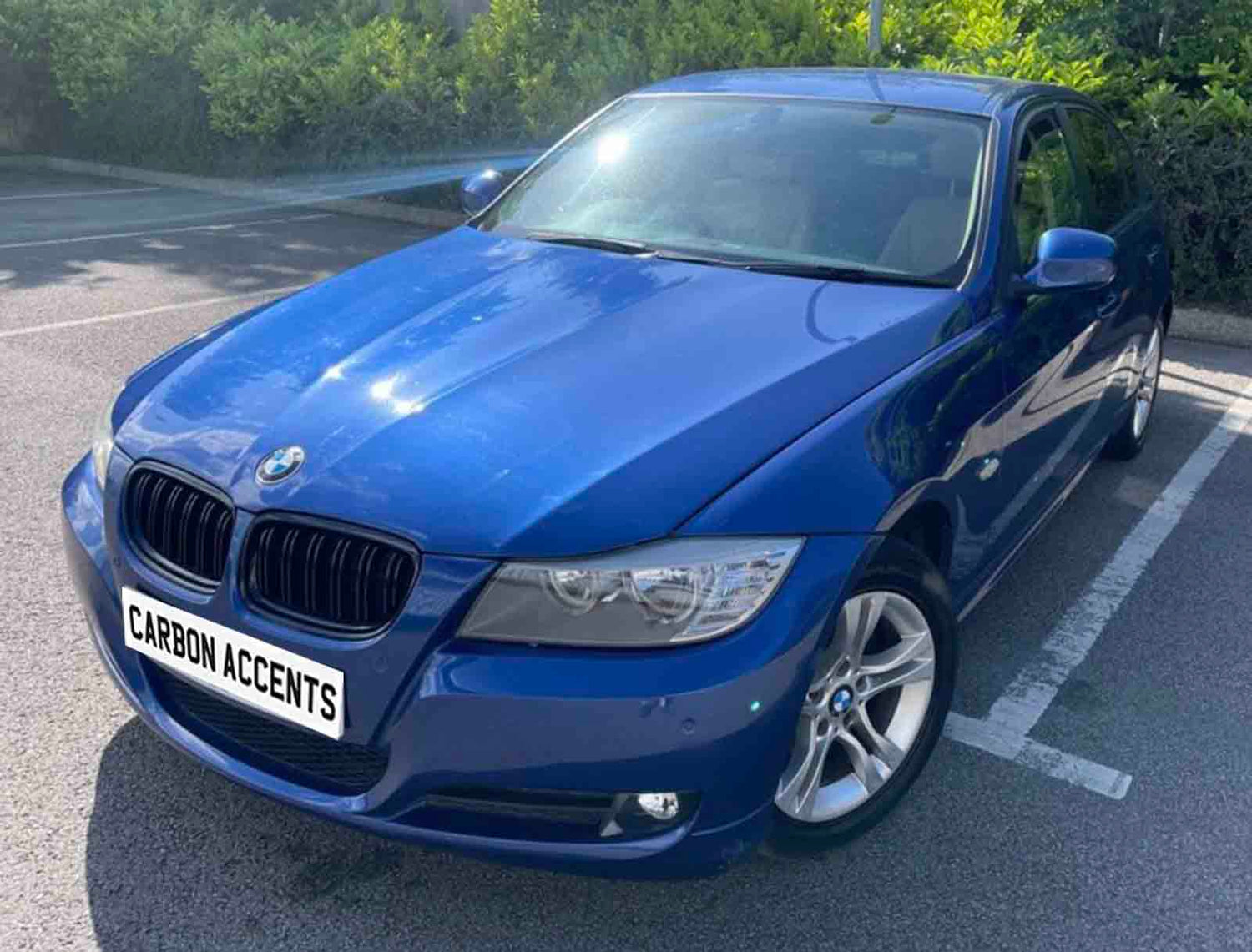 A blue car is parked in a sunlit parking space, surrounded by greenery. The license plate reads "CARBON ACCENTS."