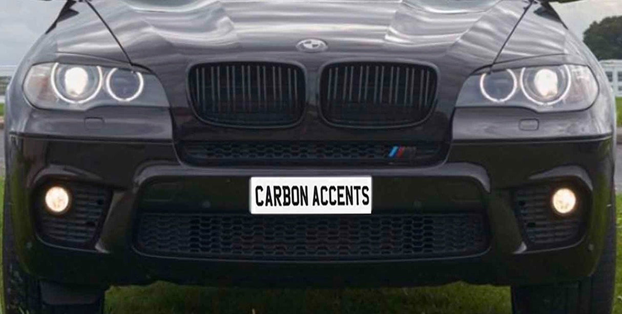 A black SUV's front view, with illuminated headlights and fog lights, is parked on grass. The license plate reads "CARBON ACCENTS."