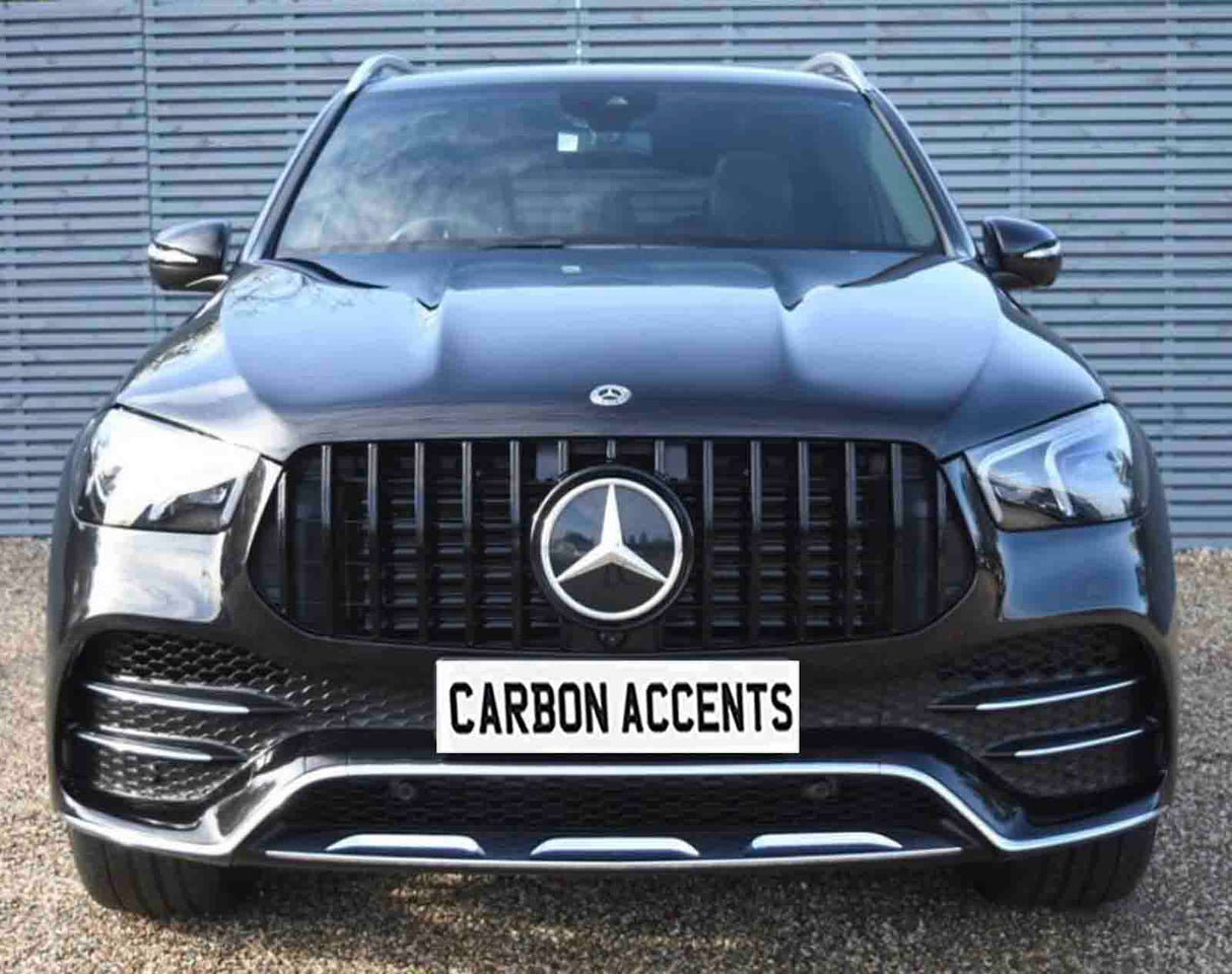 A black Mercedes-Benz SUV is parked on gravel, facing forward. It features prominent grille with a logo; the license plate reads "CARBON ACCENTS." A striped wall is in the background.
