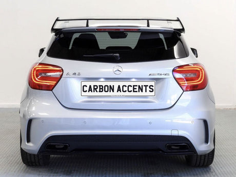 A silver hatchback car, viewed from the rear, has a prominent spoiler and dual exhaust. Text reads "CARBON ACCENTS" on the license plate. It is indoors on a patterned floor.