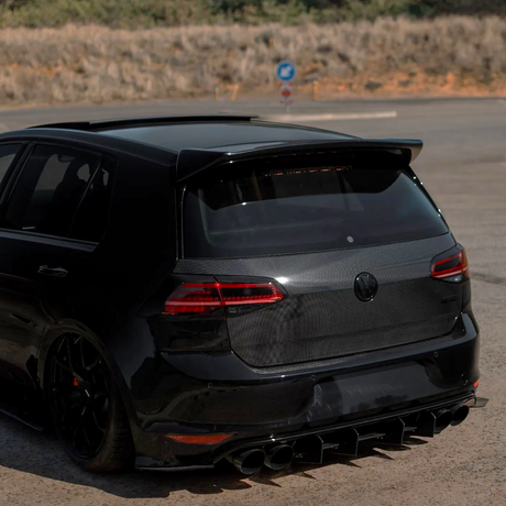 A black hatchback car is parked on a pavement with a carbon fiber rear, featuring prominent tailpipes and a roof spoiler, surrounded by a dry, grassy landscape.