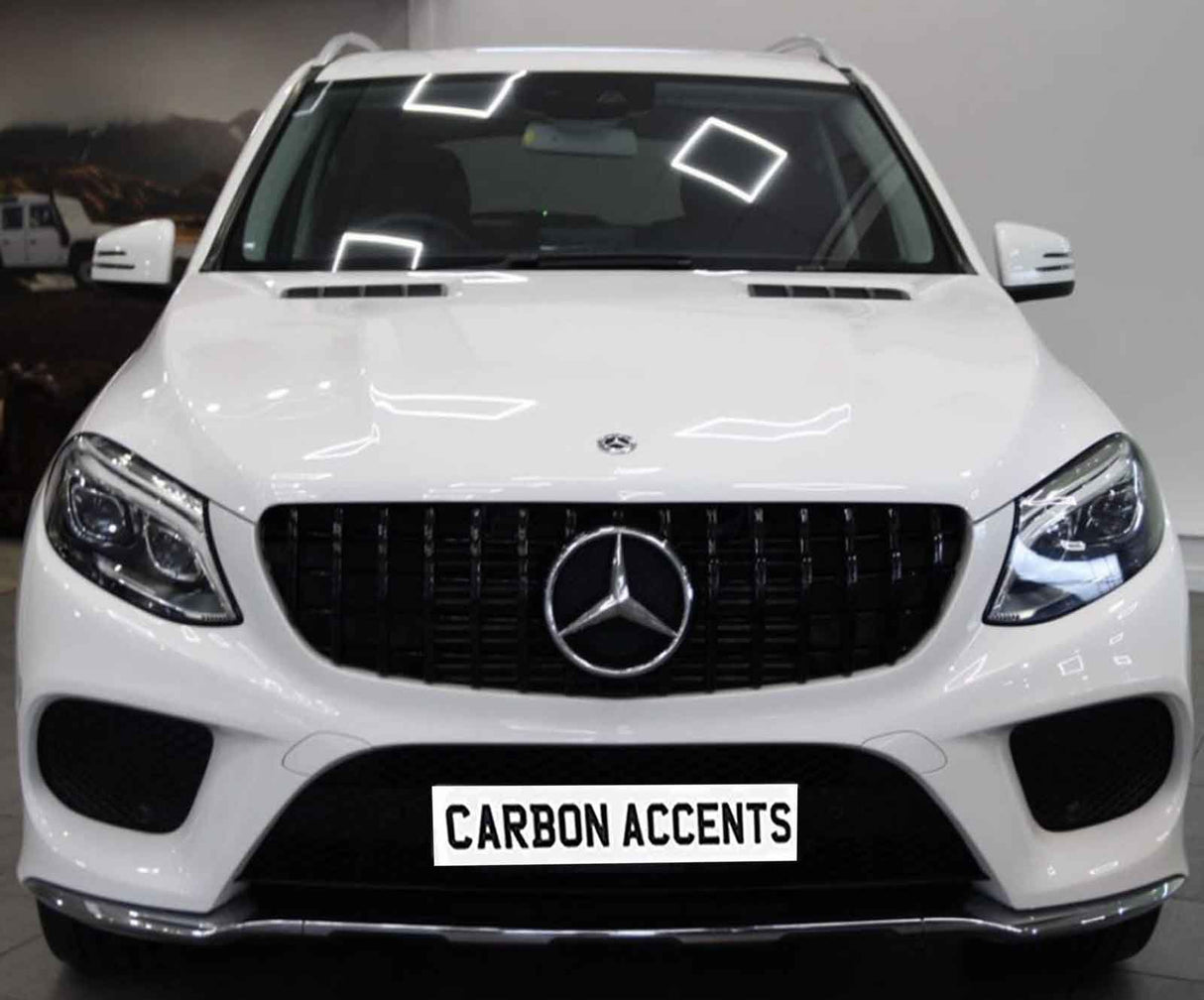 A white Mercedes-Benz car is stationary indoors, showcasing its front grille and headlights. The license plate reads "CARBON ACCENTS." Reflections of ceiling lights are visible on the vehicle's surface.