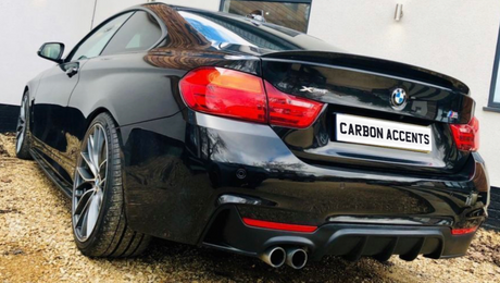 A black sports car with dual exhaust is parked on a gravel driveway beside a white building. The license plate reads "CARBON ACCENTS."
