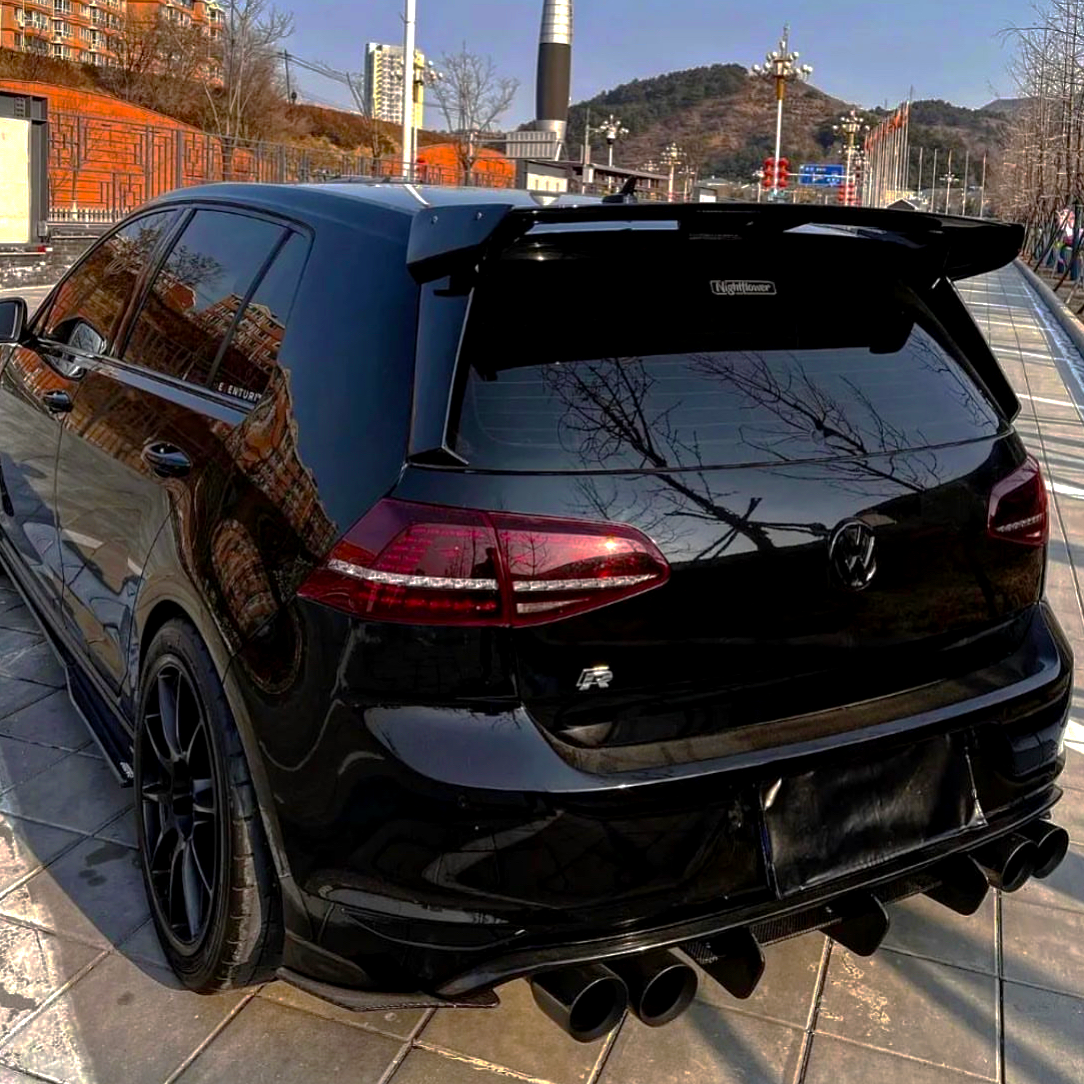 A black car, parked on a tiled pavement, features a prominent rear spoiler and quad exhaust pipes. It's set against an urban backdrop with buildings and hills in the distance.