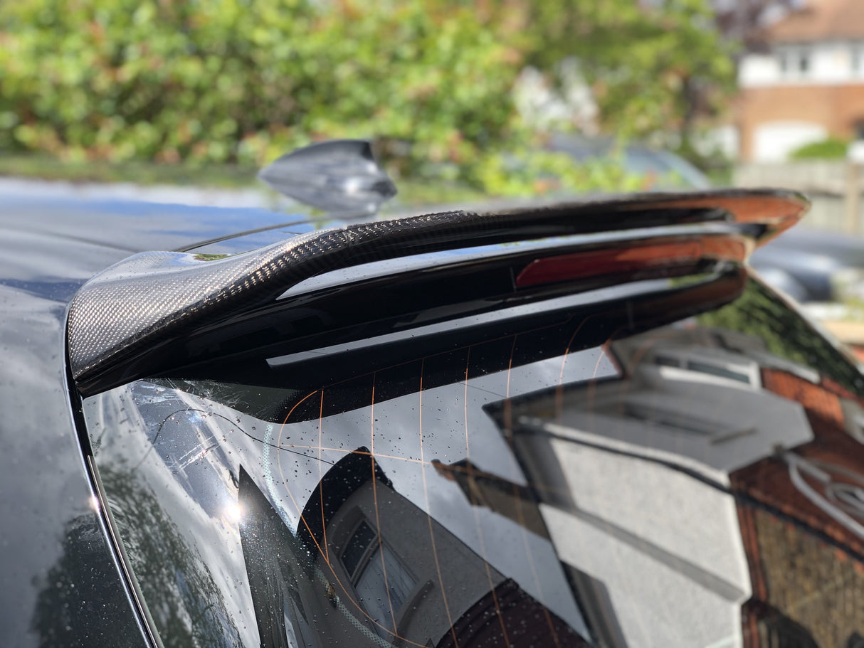 A carbon fiber car spoiler is attached to the rear of a black vehicle, glistening in sunlight. In the background, residential houses and greenery are slightly blurred.