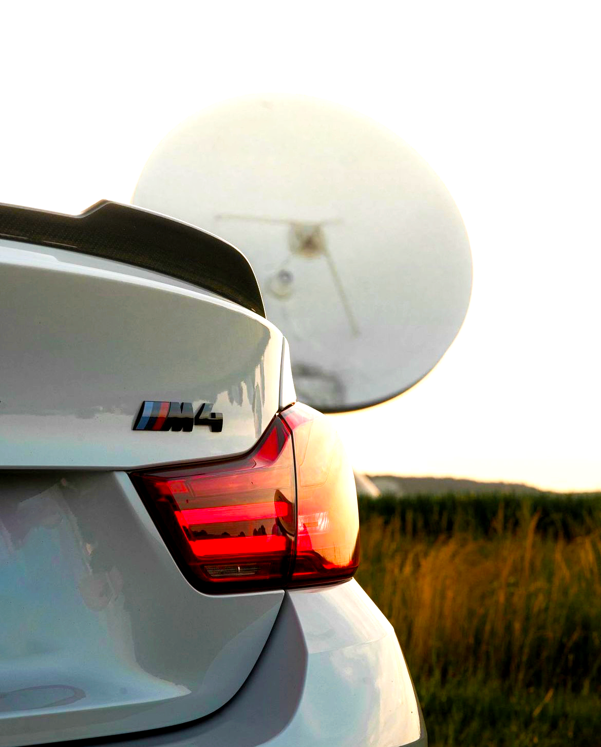 A white car with "M4" emblem and illuminated tail light is parked, partially obscuring a large satellite dish in a grassy field at sunset.