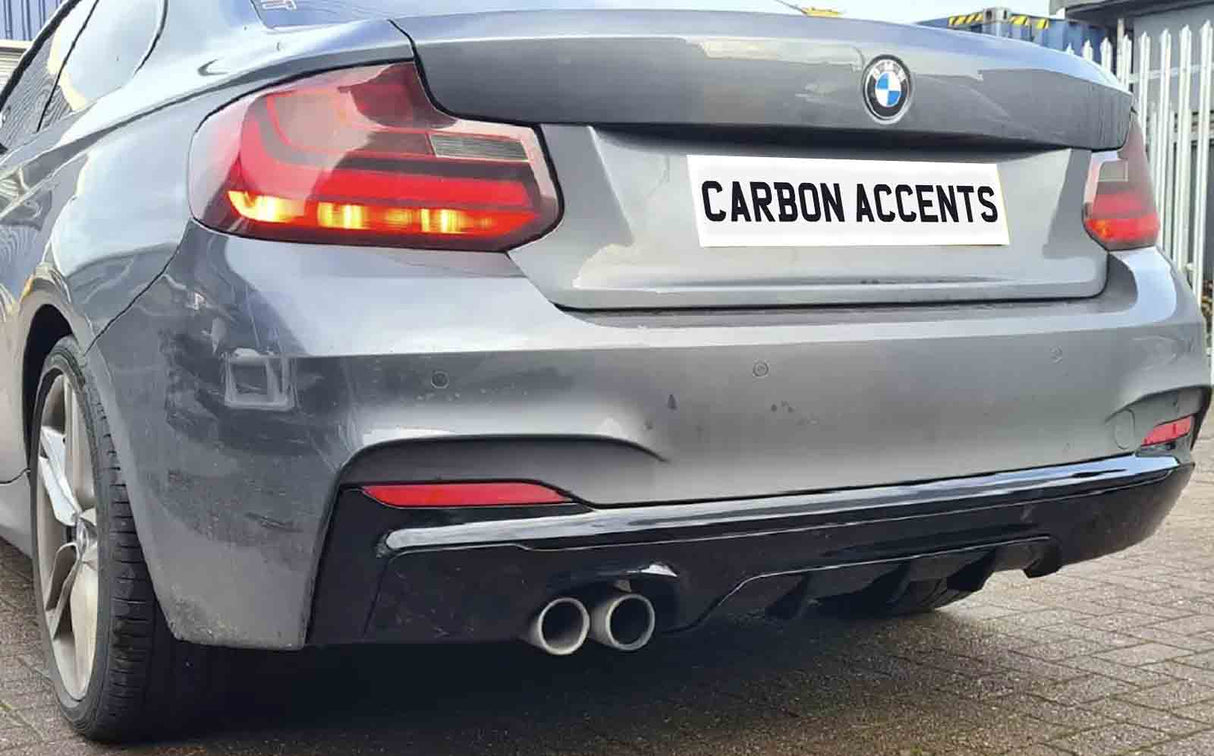 A gray BMW car is parked, displaying dual exhausts below a license plate reading "CARBON ACCENTS." It's situated on a paved surface, adjacent to a metal fence.